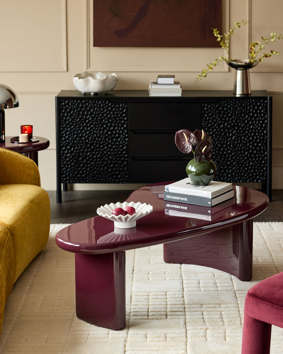 Gloss plum coffee table with curved oval top and sculptural pedestal base, styled in modern living room setting.