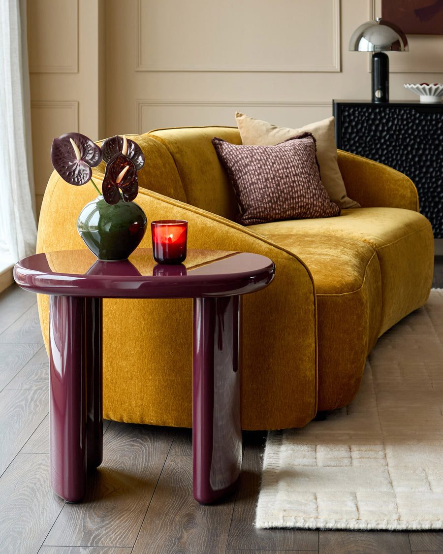 Gloss plum side table with curved, organic shape, styled next to a mustard yellow sofa in a modern living room setting.