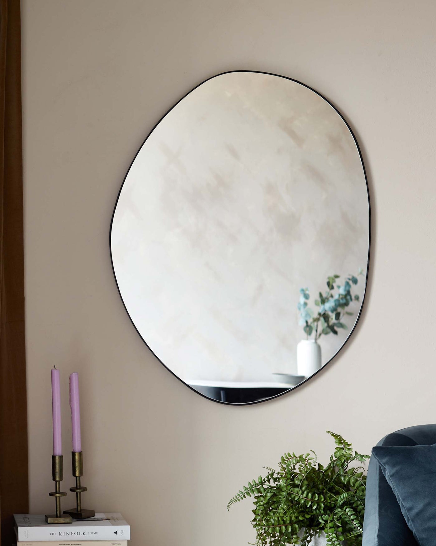 Elegant oval wall mirror with a slender black frame, alongside a small minimalist side table featuring a stack of books, a brass dual candlestick holder with lavender candles, and a simple white vase with eucalyptus branches.