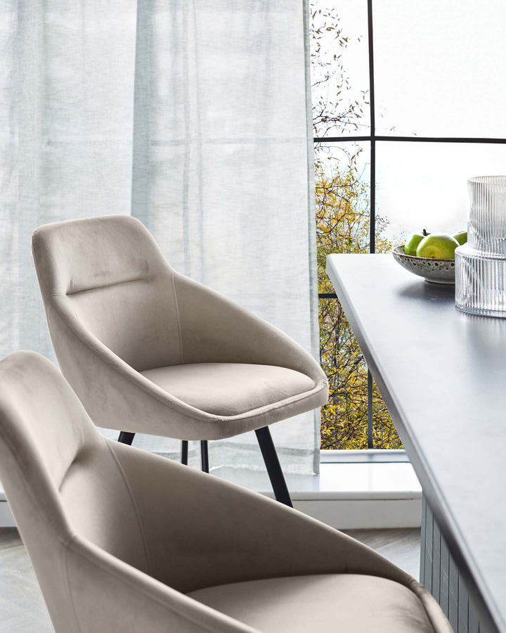 Beige upholstered chairs sit beside a table with fruit and glassware. A large window and sheer curtain provide natural light.