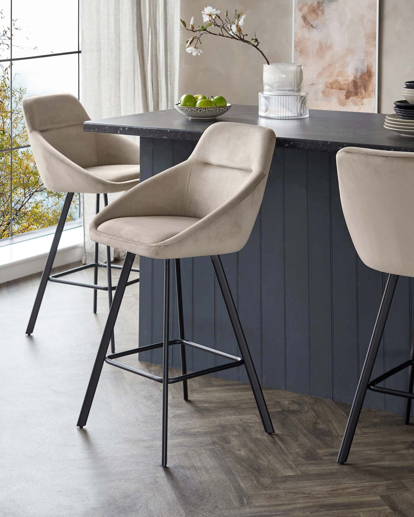 Elegant modern bar stools with beige upholstery and black metal legs, paired with a dark stone countertop kitchen island in a stylish interior with a herringbone wood floor.