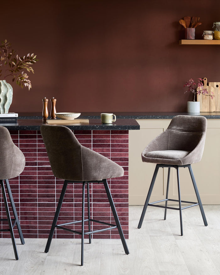 Three stylish, grey, fabric bar stools with black metal frames stand at a kitchen island. Kitchenware is displayed on the counter.