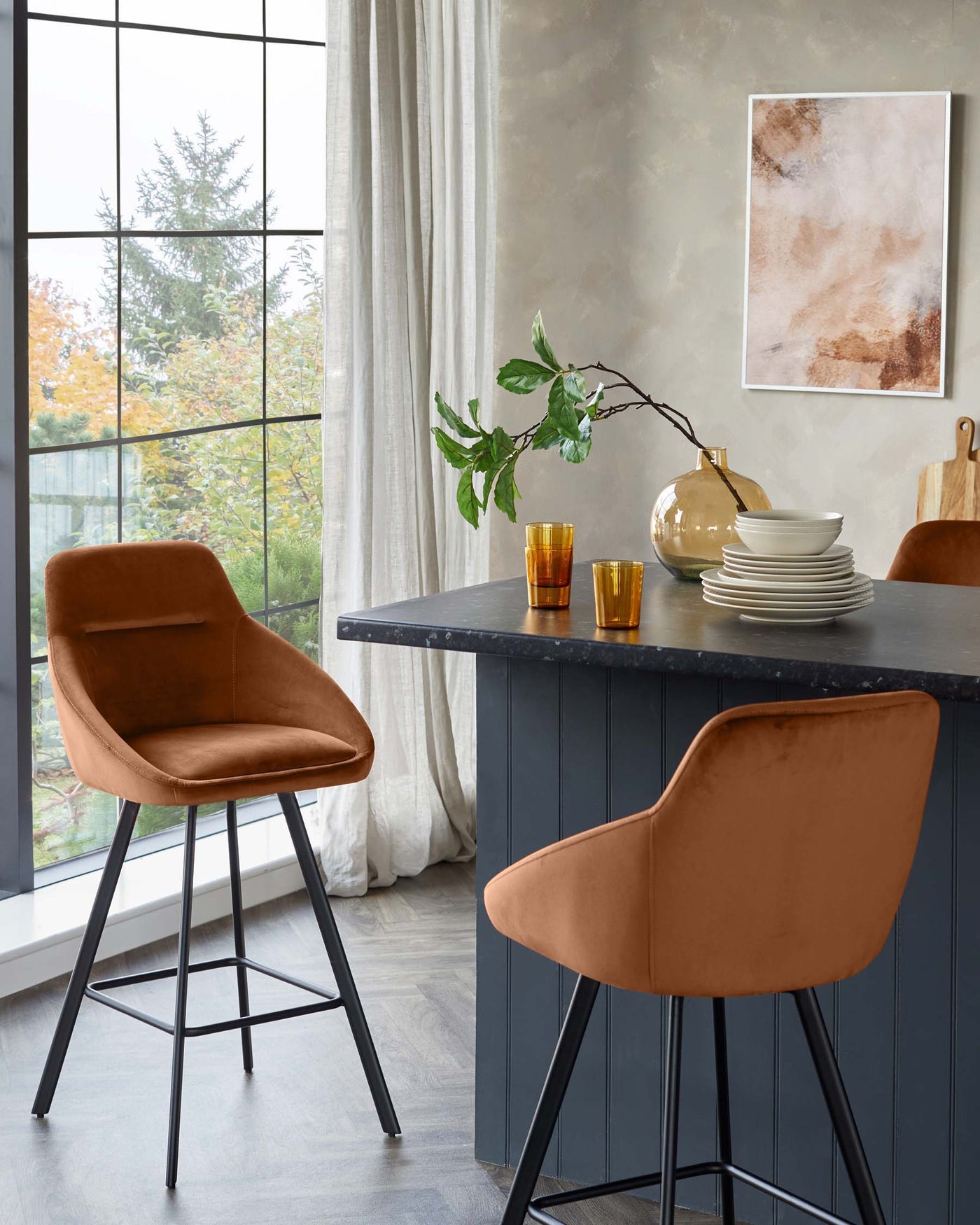 Elegant contemporary bar stools with plush, velvet upholstered seating in a rich terracotta colour, featuring a curved backrest and mounted on sleek, black metal legs with footrests, complementing a stylish black kitchen island with a textured finish.