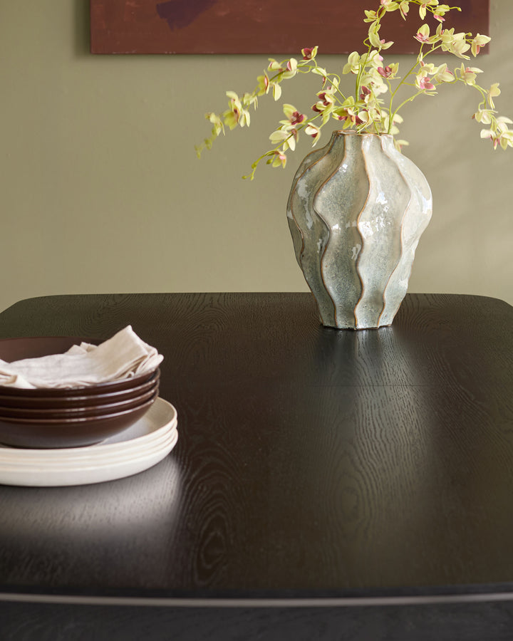 Black wood extending dining table with wavy edge detail, styled with stacked plates and floral arrangement.