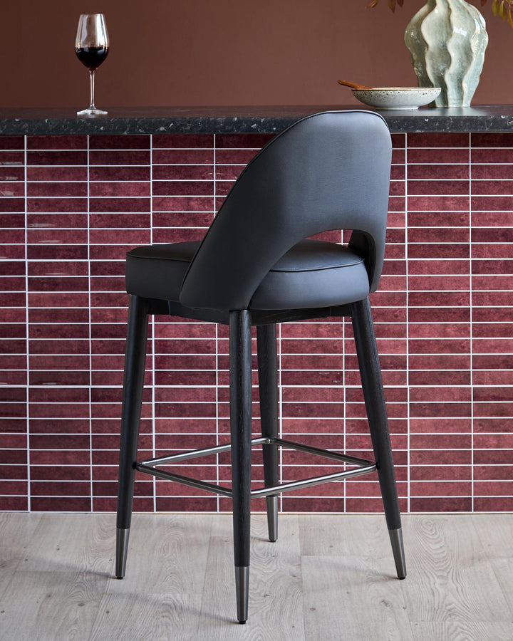 A stylish black bar stool with a leather seat and a metal footrest, sits in front of a brick-tiled wall.