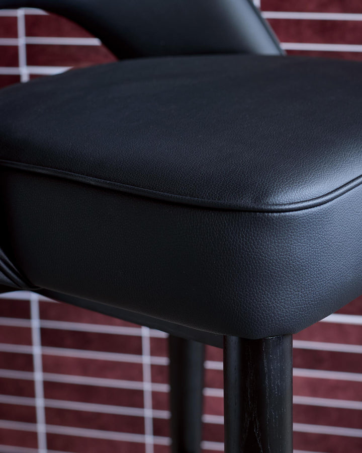 Close up of a black padded bar stool with a dark wood frame against a red brick background.
