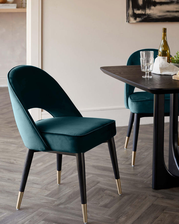 Two stylish teal velvet dining chairs with black legs and brass accents next to a modern, dark wood table.