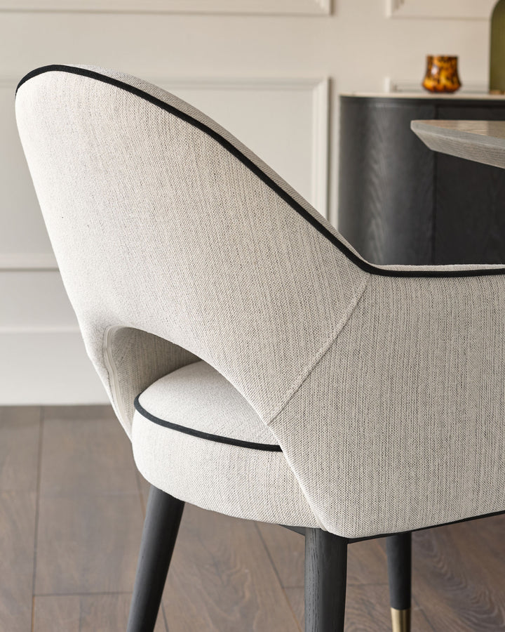 Clover dining armchair in ivory fabric, side angle view showing curved backrest with black piping detail and dark wooden legs.