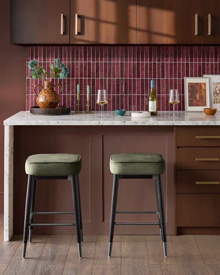 Clover velvet backless bar stool in olive green with dark wooden legs, shown in a kitchen setting.