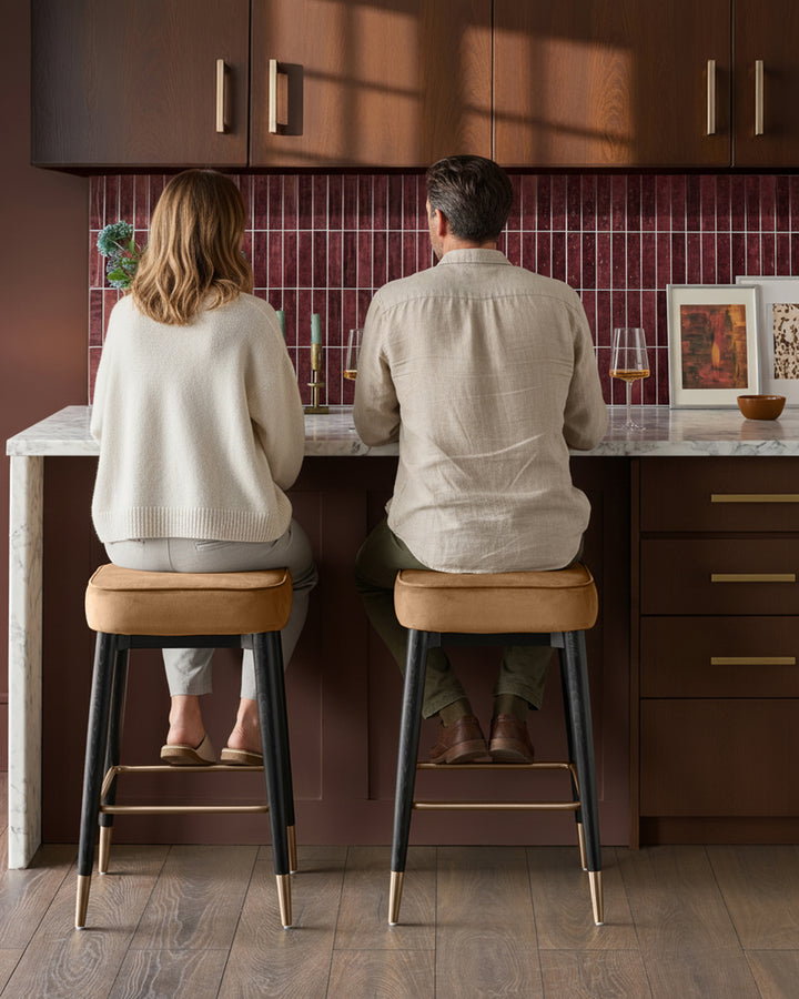 Caramel fabric backless bar stools with black legs and brass footrests, shown at kitchen counter.