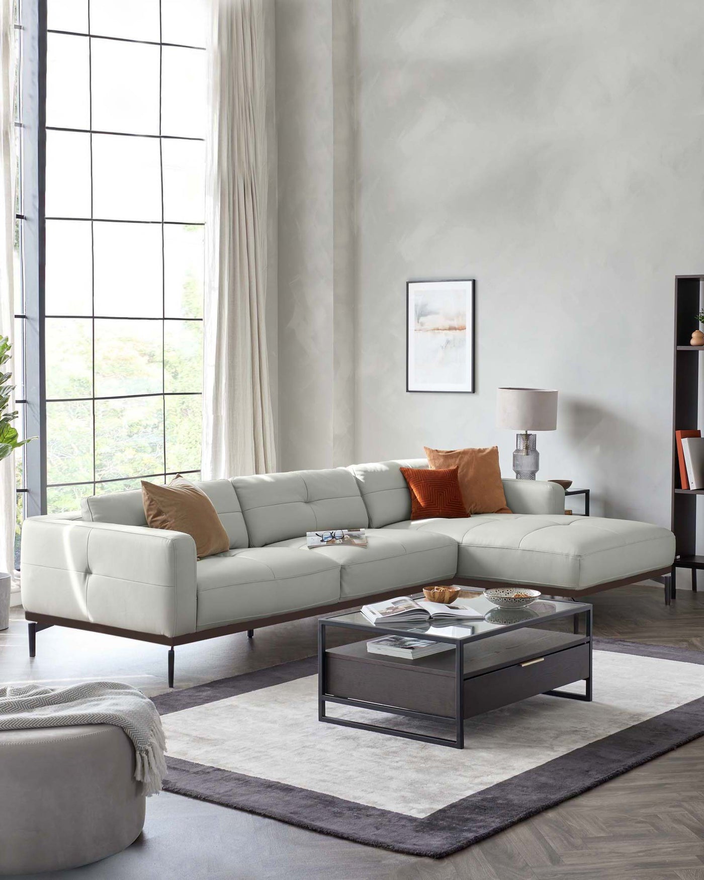Elegant light grey L-shaped sectional sofa with plush cushioning and throw pillows in earth tones, paired with a modern rectangular coffee table with a black metal frame and dark wood top. The ensemble is accented by a soft grey ottoman, all arranged on a two-tone area rug over a herringbone-patterned floor.