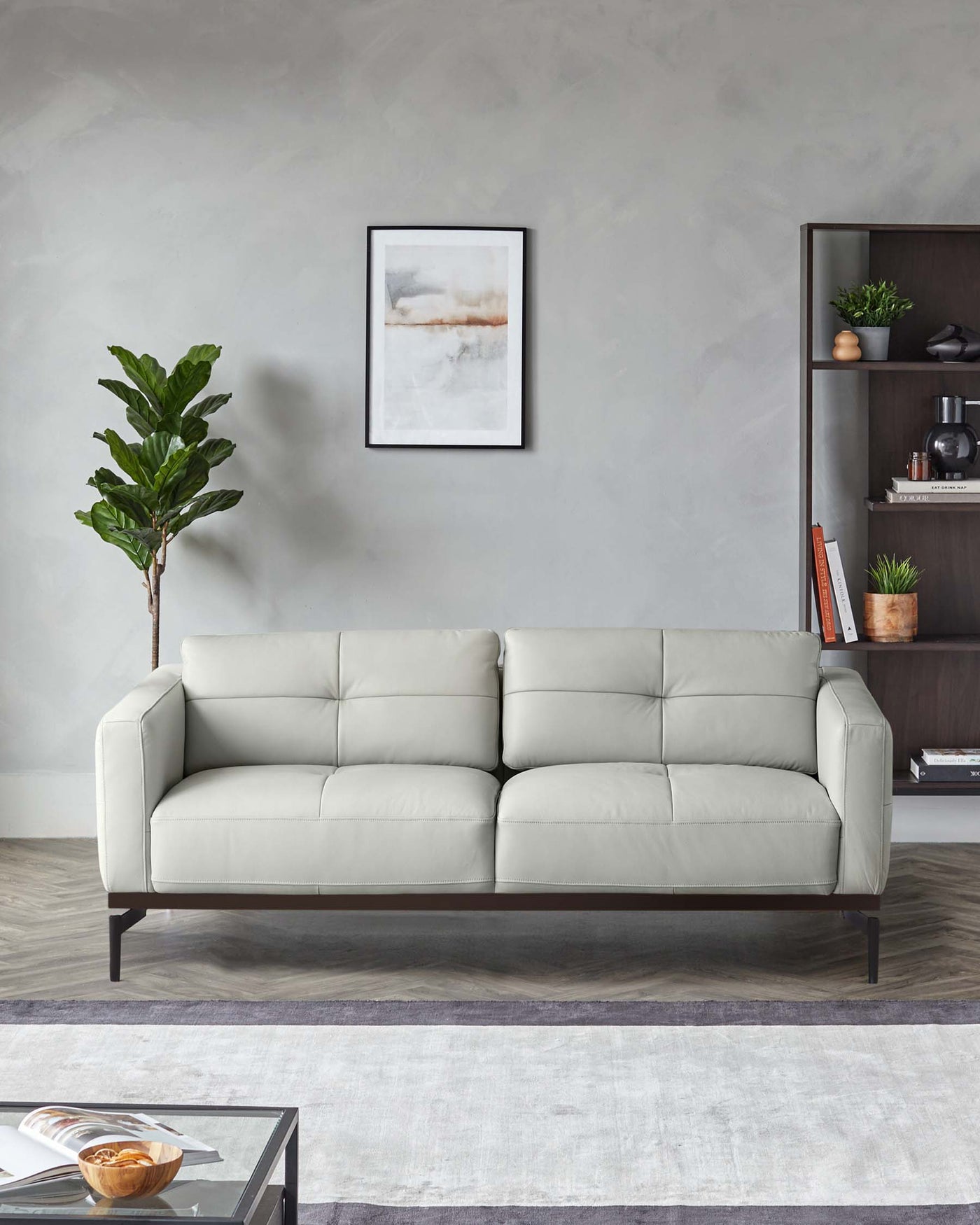 A modern light grey three-seater sofa with clean lines, tufted seat cushions, and dark wooden legs, paired with a dark brown wooden bookshelf filled with decorative items and books, set against a neutral-toned living room backdrop.