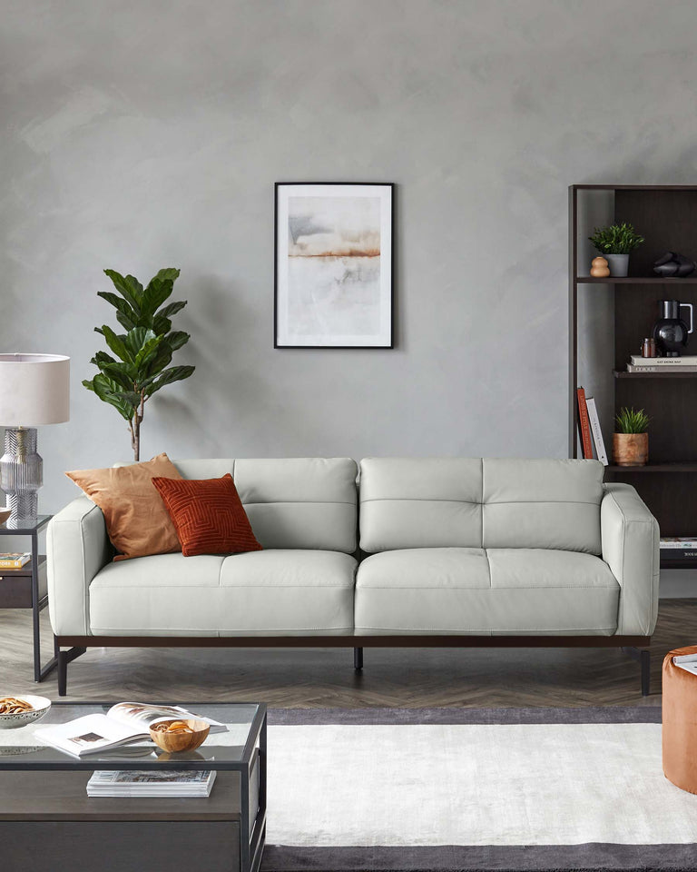 A contemporary light grey three-seater leather sofa with clean lines and dark tapered legs, accompanied by a modern dark wood side table and a coordinating coffee table. Accessories include a white and grey area rug, decorative pillows, and a potted indoor plant adding a touch of greenery to the space. A framed abstract artwork hangs on the wall above the sofa, and a shelf with books and small plants contributes to the modern aesthetic.