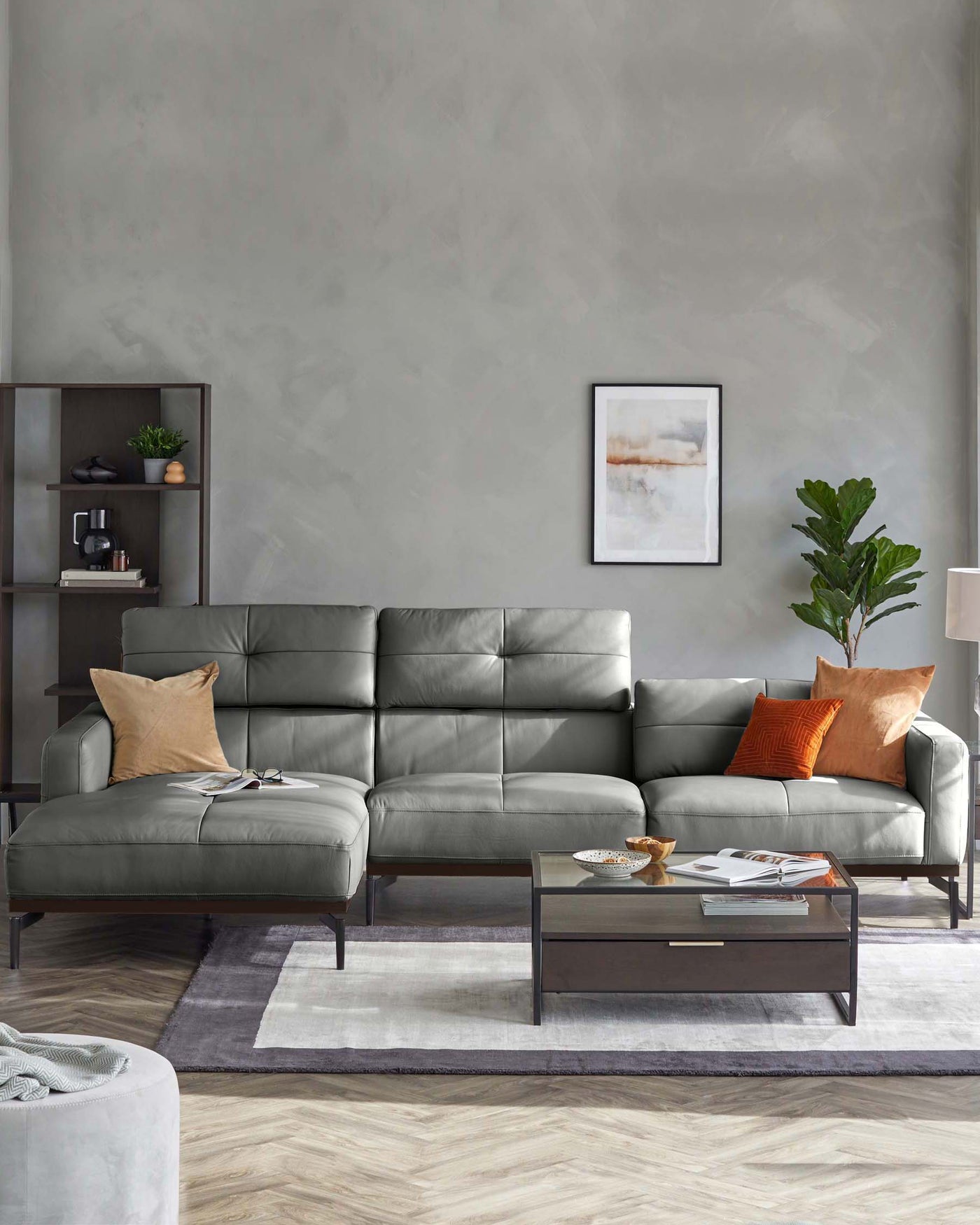 Modern living room featuring a sleek grey L-shaped sectional sofa adorned with beige and orange pillows, complemented by a rectangular wooden coffee table with a glass top. To the side stands a minimalist dark-wood shelving unit displaying decorative items. A framed abstract wall art adds an artistic touch above the couch, and a potted plant brings a splash of greenery to the space. The area is anchored by a two-tone area rug over a herringbone patterned floor.