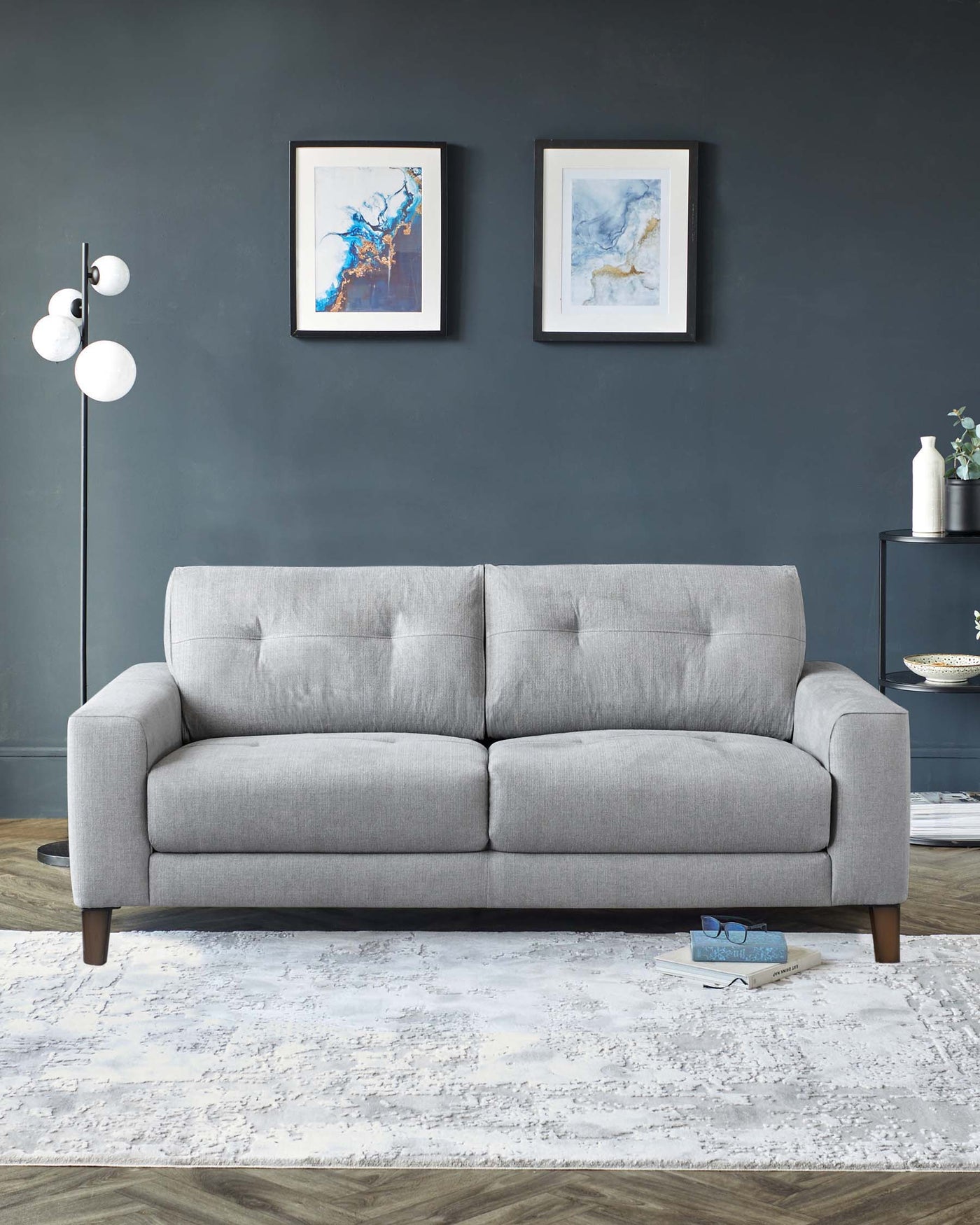 Modern three-seater sofa with tailored lines and dark wooden legs, upholstered in light grey fabric. Positioned on a white and grey patterned area rug, set against a dark grey wall featuring abstract framed artwork.