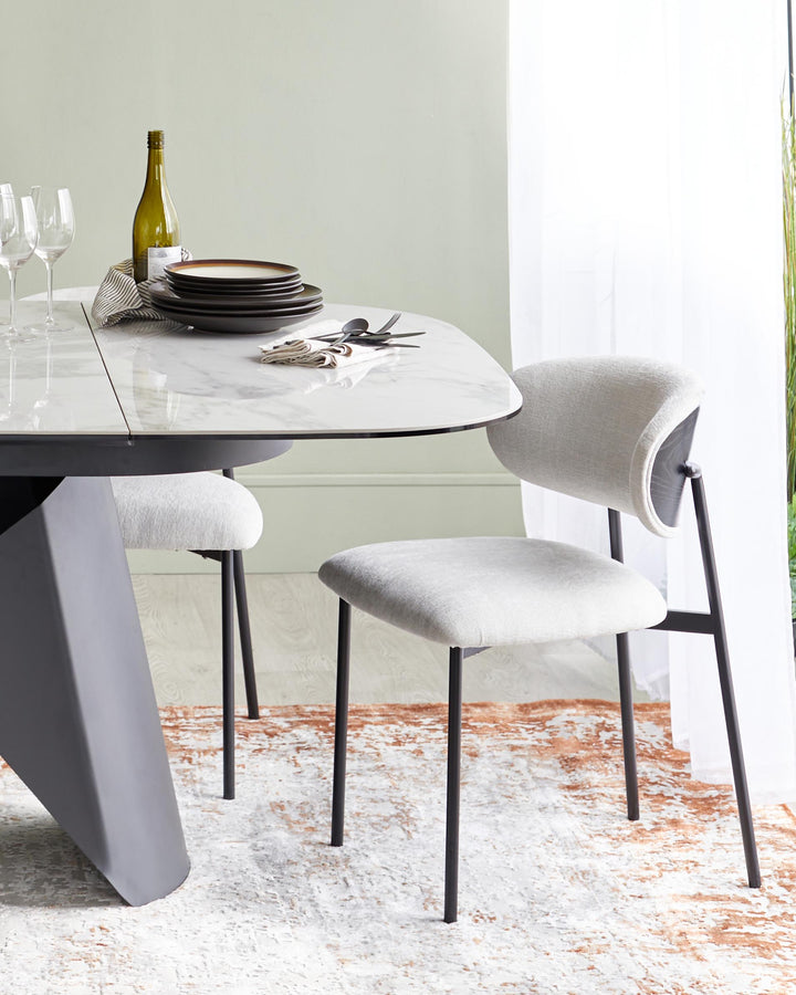 A modern dining table with a marble top and geometric base, paired with sleek grey upholstered chairs on a textured rug.