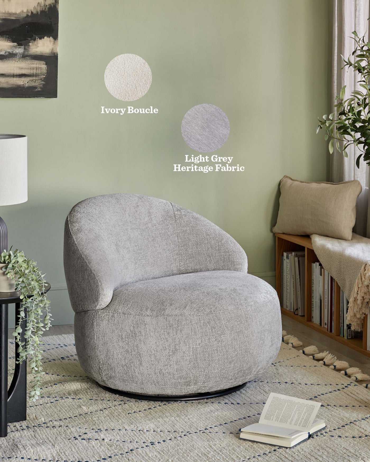 Contemporary light grey fabric upholstered armchair with a rounded, organic design, featuring a raised backrest and a wide, comfortable seat. The chair is positioned on a textured area rug with a neutral palette accented with blue lines, alongside a small side table with cascading greenery. In the background, a cosy nook is seen with a tan cushioned reading chair and a stack of books on a wooden shelf, enhancing the tranquil and inviting ambiance of the space.