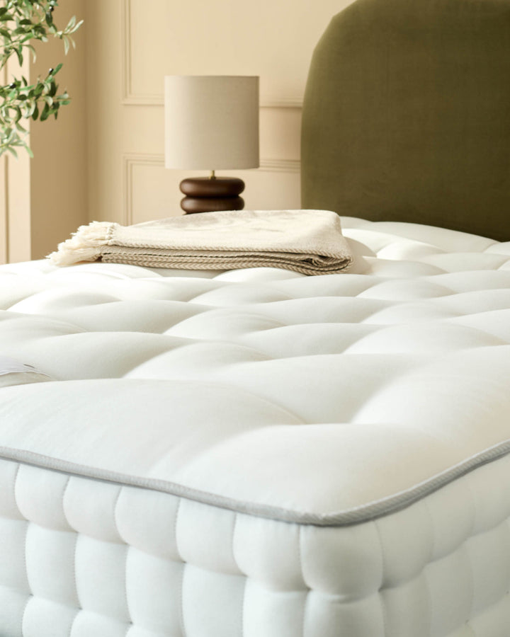 Close-up of a white, quilted mattress with a green headboard, topped with a folded beige blanket and a lamp in the background.