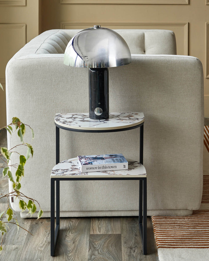 A stylish two-tier marble effect side table with a chrome lamp and book, set against a neutral sofa.
