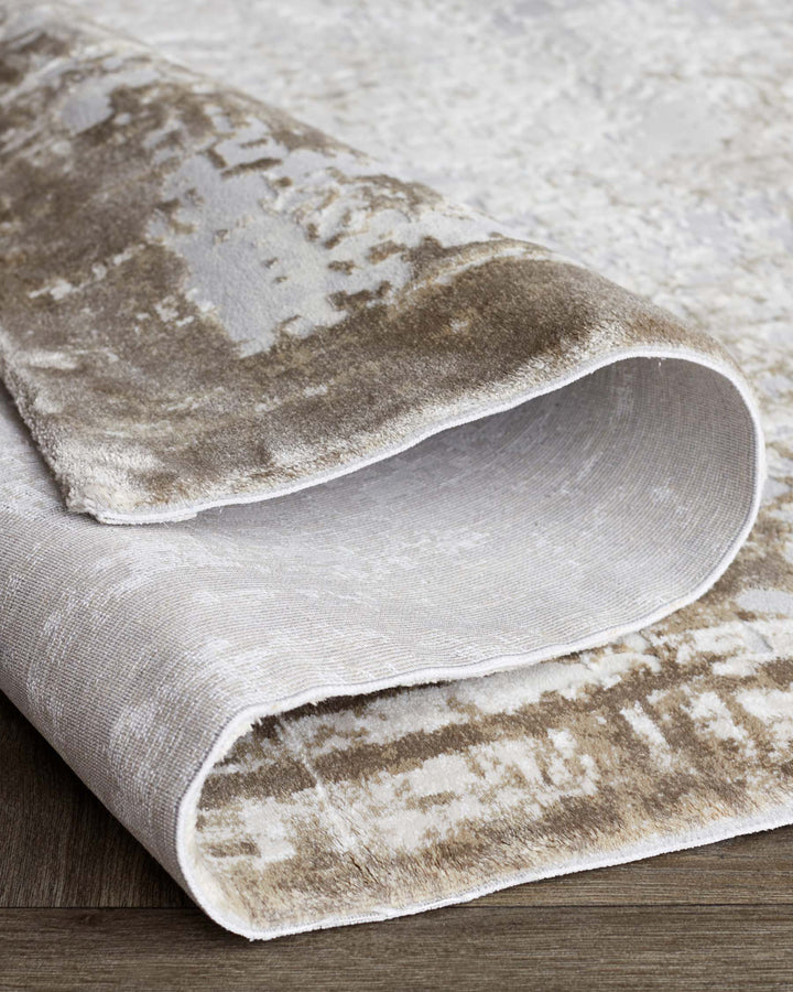 Elegant beige and brown area rug with a plush texture and abstract pattern, partly rolled up on a wooden floor.