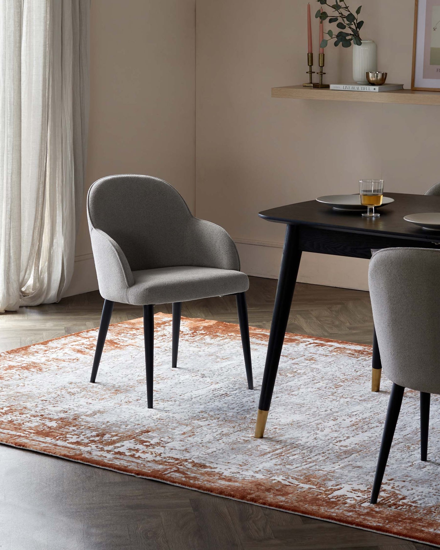 Modern dining setting featuring a round black table with an elegantly textured surface, complemented by a set of sophisticated grey upholstered chairs. Each chair exhibits a gently curved backrest and seat, supported by angled black legs accented with gold-toned tips, harmoniously blending comfort and luxury. The arrangement is centred on a distressed white and terra cotta area rug, which adds a warm, inviting touch to the scene.