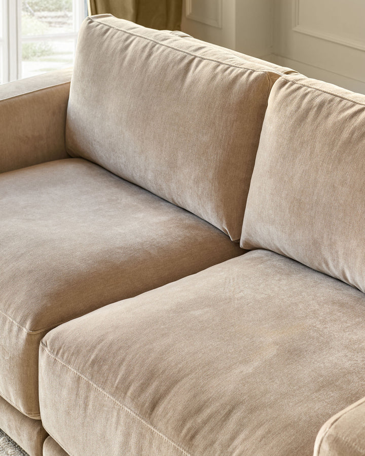 Eden 2-seater sofa in champagne fabric, angled view showing cushioned seat, back pillows, and rolled arms near a window.