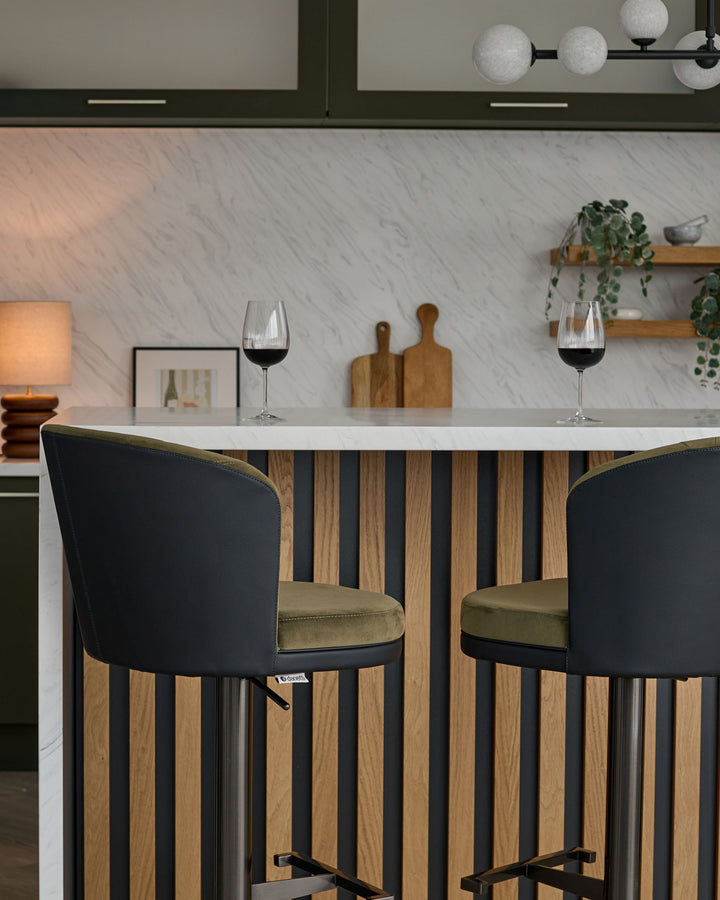 Elodie gas lift bar stool in black velvet with olive-green seat, shown at modern kitchen counter with striped wood base.