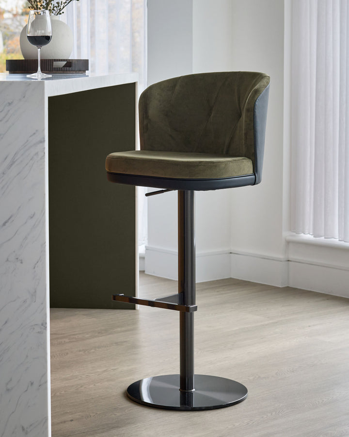 Elodie gas-lift bar stool in olive green velvet with black metal base and footrest, shown at kitchen counter.