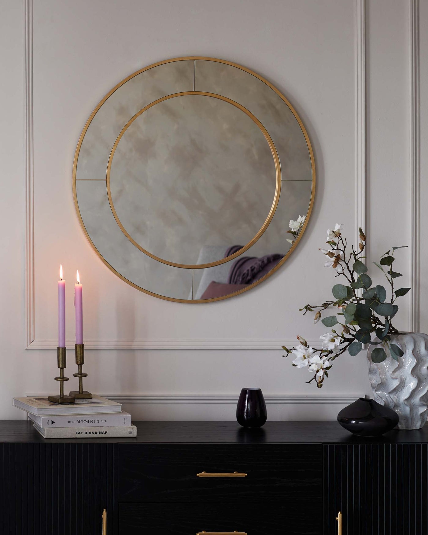 Elegant modern black sideboard cabinet with vertical ridges and brass handles, topped with minimalist decorative items including a brass two-candle holder with lit candles, stacked coffee table books, and assorted vases, set against a wall with a large round mirror featuring a cloudy reflective surface and a thin gold frame.