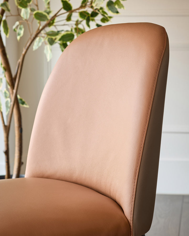 Tan leather Emerson dining chair shown in close-up detail of the backrest and seat, with decorative plant visible behind.