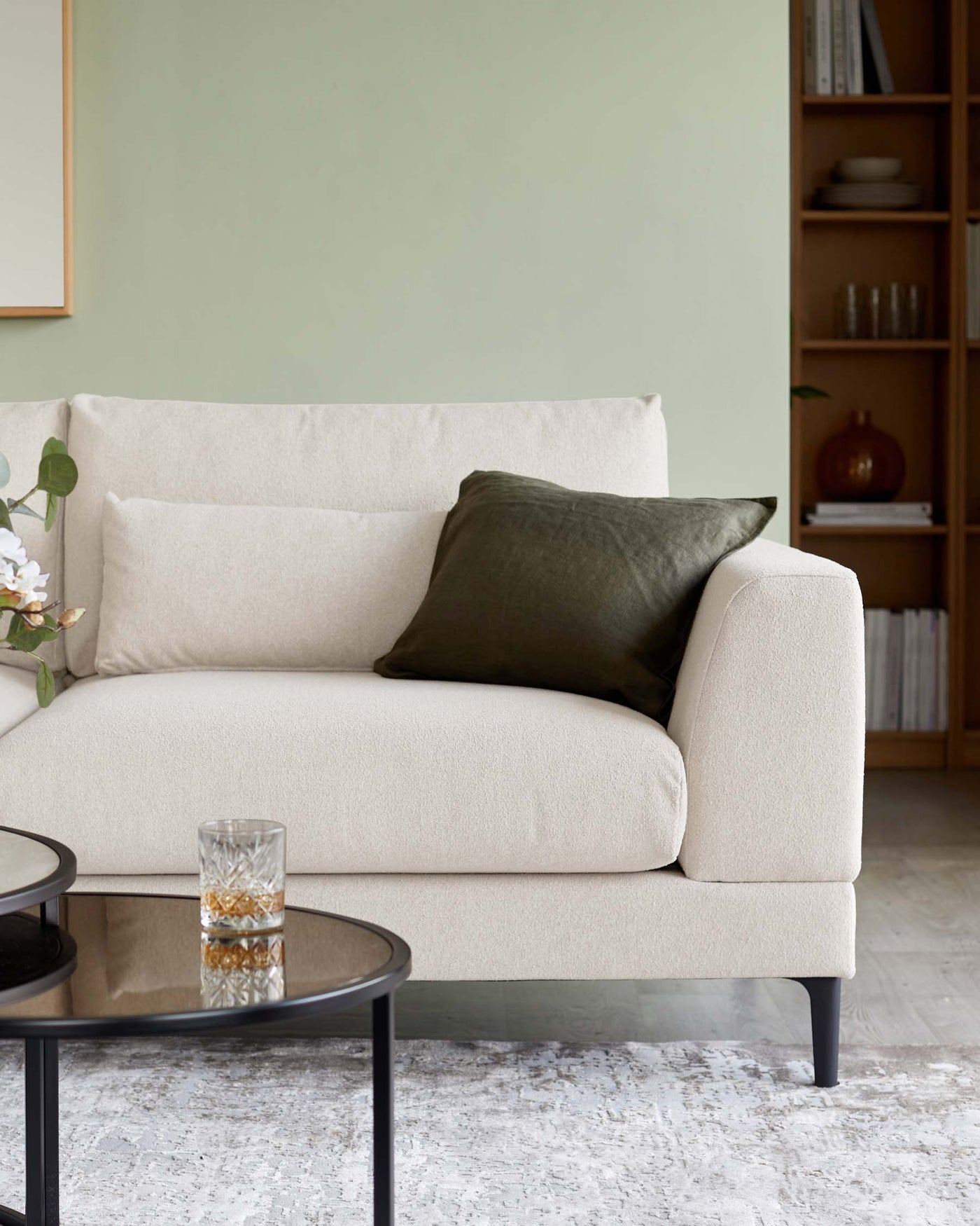 A contemporary beige three-seater sofa with clean lines and minimalistic design, featuring cushioned seats and backrest, and accompanied by a dark olive rectangular lumbar pillow. The sofa is positioned next to a round, black-framed coffee table with a glass top, all resting on a textured off-white area rug. In the background, there's a tall wooden bookshelf partially filled with books and decorative items against a light green wall.