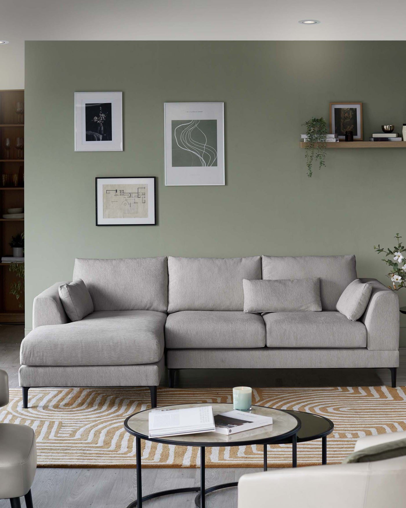 Modern living room with a light grey fabric L-shaped sectional sofa featuring plush back cushions and a chaise lounge on the left. In front of the sofa, there is a round, two-tiered coffee table with a black metal frame and glass tops. A beige and white patterned rug is beneath the coffee table and sofa. On the left, part of a light-coloured armchair is visible. The room is accessorized with framed wall art and a wooden wall shelf with decorative items.