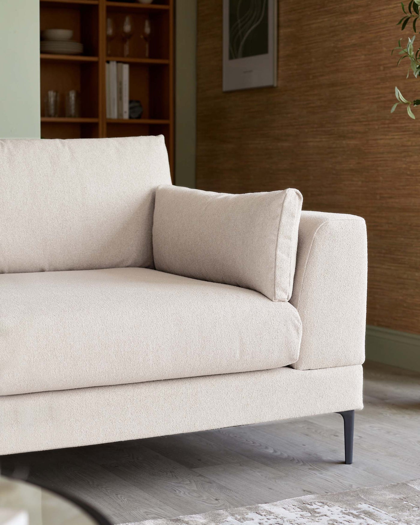 A contemporary beige upholstered sofa with clean lines and a minimalist design. Features plush cushioning, a single bench-style seat cushion, and a structured backrest with an additional loose pillow for comfort. Rests on slim, black metal legs complementing its modern aesthetic. Background includes a wood cabinet with glassware and books, set against a textured wall with framed artwork, enhancing the sophisticated ambiance.