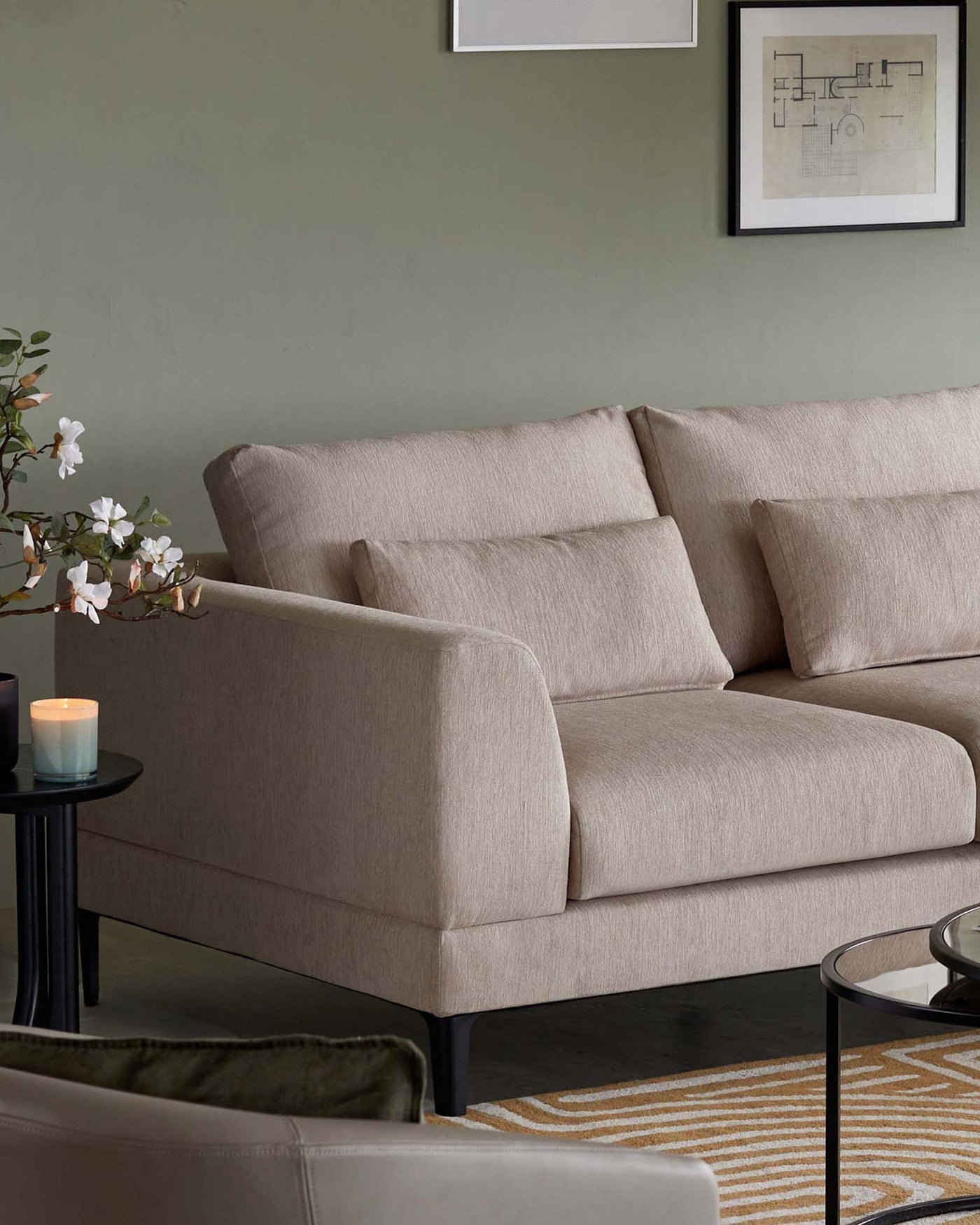 A contemporary beige three-seater sofa with a minimalist design, featuring straight lines and square-edged cushion padding. The sofa sits on low-profile dark legs, complementing a sleek silhouette. Beside it is a small round black side table with a glass top, hosting a lit candle. Behind the sofa, there is a simple artwork in a black frame, enhancing the modern aesthetic of the room. A cream-colored carpet with yellow and beige geometric patterns lies partially visible on the floor.