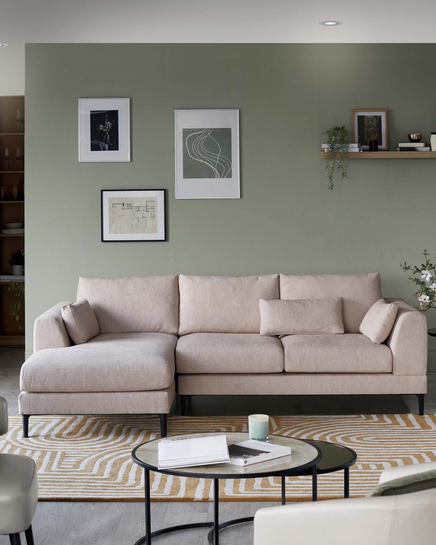 Elegant beige fabric L-shaped sectional sofa with plush cushions and a simple, modern design. In front of the sofa is a round, black metal coffee table with a glass top, hosting books and a candle. The furniture is complemented by a patterned off-white and beige area rug beneath.