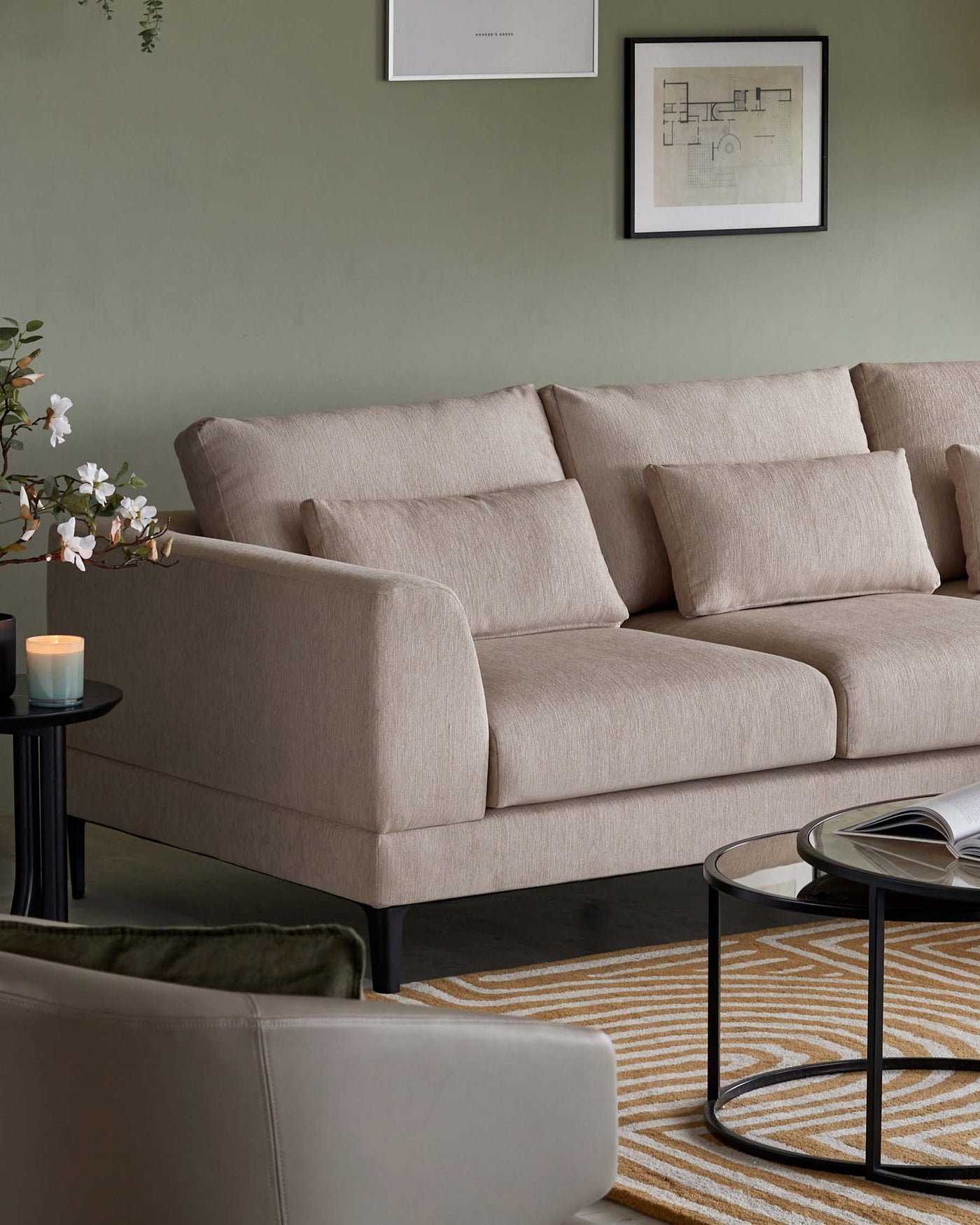 Beige fabric three-seater sofa with plush back cushions and minimalist black legs, paired with a round glass-top side table featuring a black frame. The sofa is complemented by a neutral-toned area rug with geometric patterns.