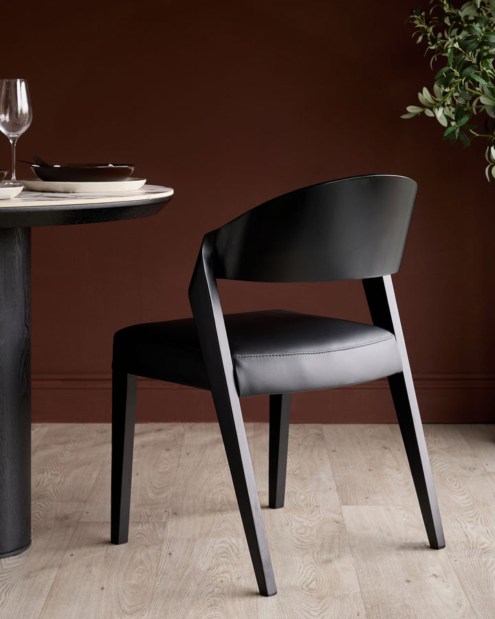 Enzo dining chair in black leather with curved backrest and wooden legs, positioned at a round table setting.