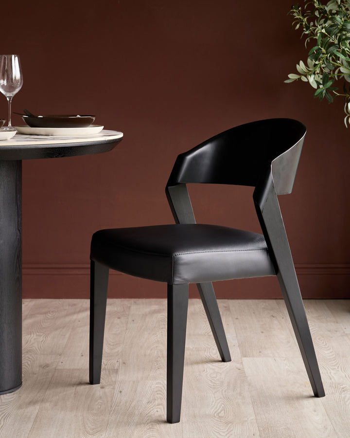 Enzo dining chair in black leather with dark wood frame, shown at round table setting with place setting and greenery.