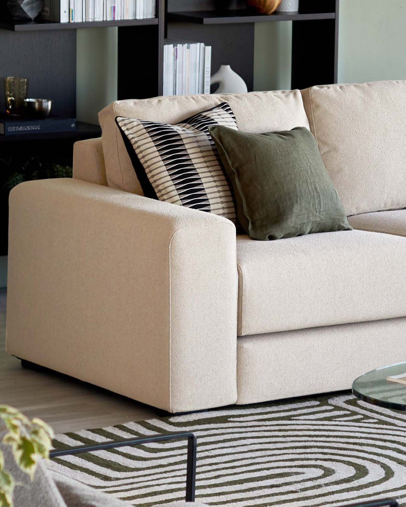 Beige upholstered sofa with a modern design, featuring clean lines, a squared-off armrest, and plush back cushions. The sofa is accented with an assortment of decorative pillows, including one with black and white stripes and another in solid olive green. In the background, a dark grey shelving unit houses an array of books and decorative objects. A round glass-top coffee table with a slim black metal frame is partially visible, all resting on a geometric-patterned area rug in muted tones.