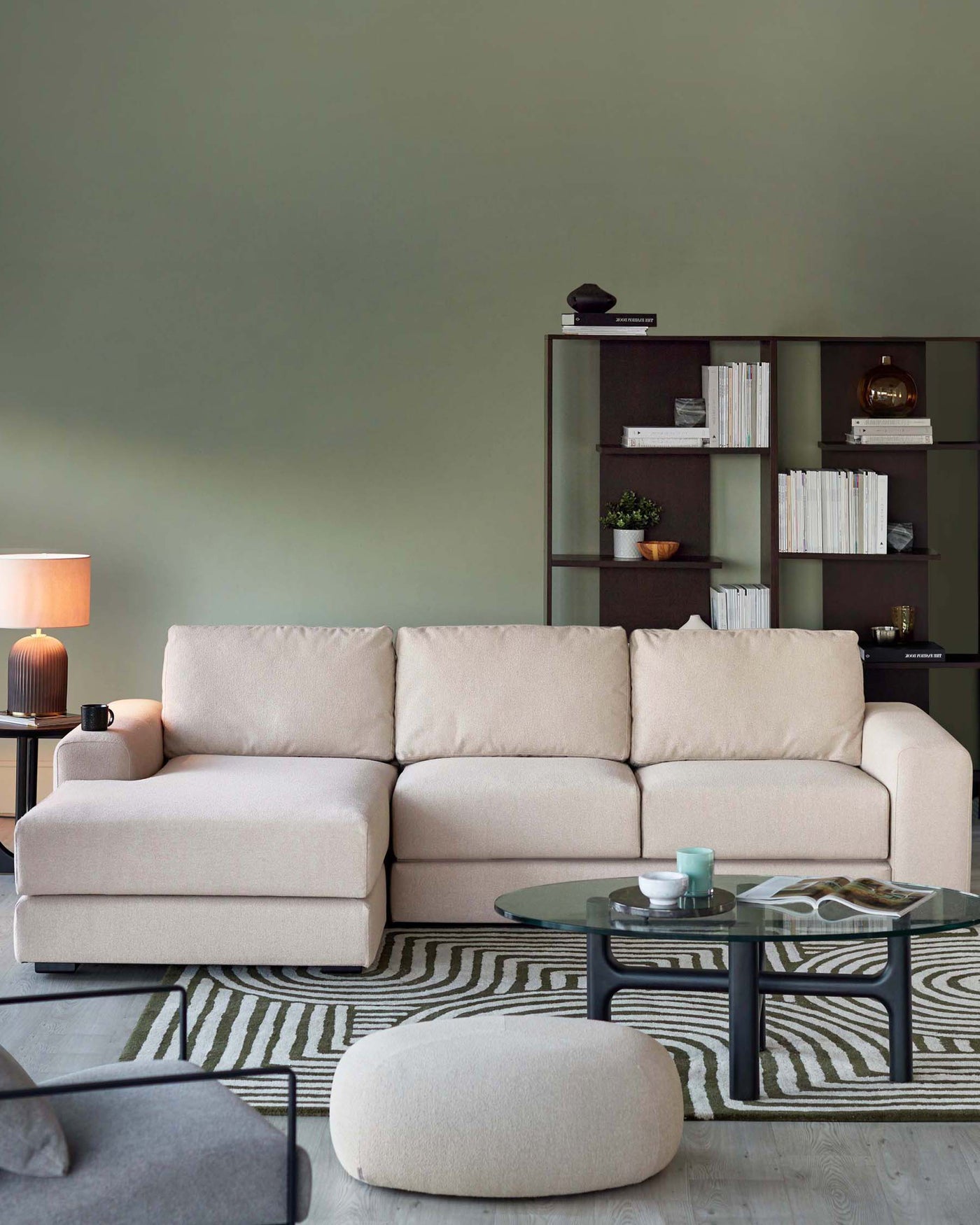 A modern living room setup featuring a light beige L-shaped sectional sofa, a round beige ottoman, a glass coffee table with a black frame, a dark bookshelf filled with white books and decorative items, and a table lamp with a beige shade on a small side table.