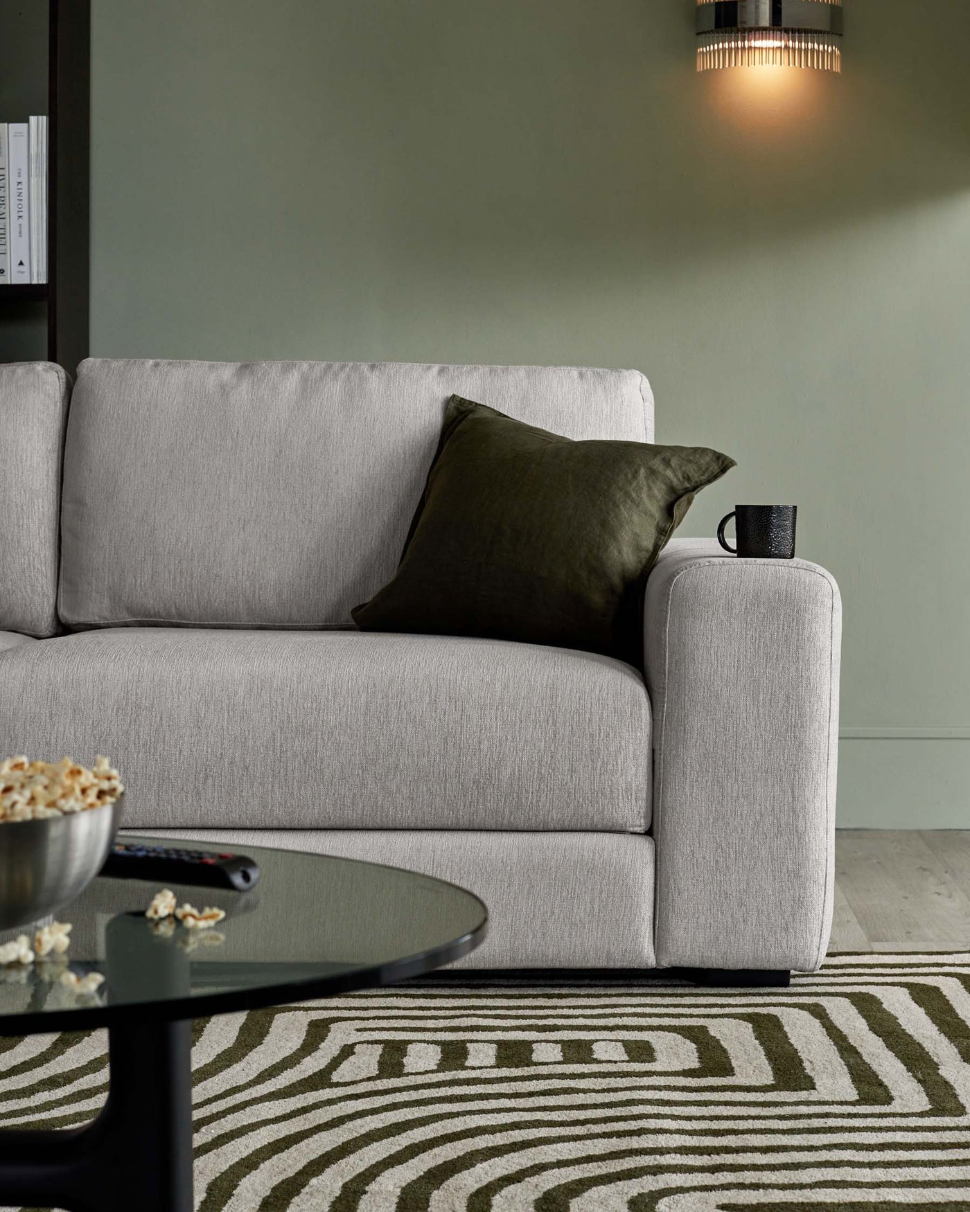 Contemporary light grey fabric sofa with clean lines and subtle texture. Features plush cushions, one dark olive green decorative pillow, and a flat, wide armrest on the left side. In front of it, a round, black matte coffee table with a smoked glass top, hosting a metallic bowl with popcorn and a remote control, placed over a patterned cream and olive area rug. Behind the sofa, a modern wall lamp with a cylindrical shade provides soft lighting.