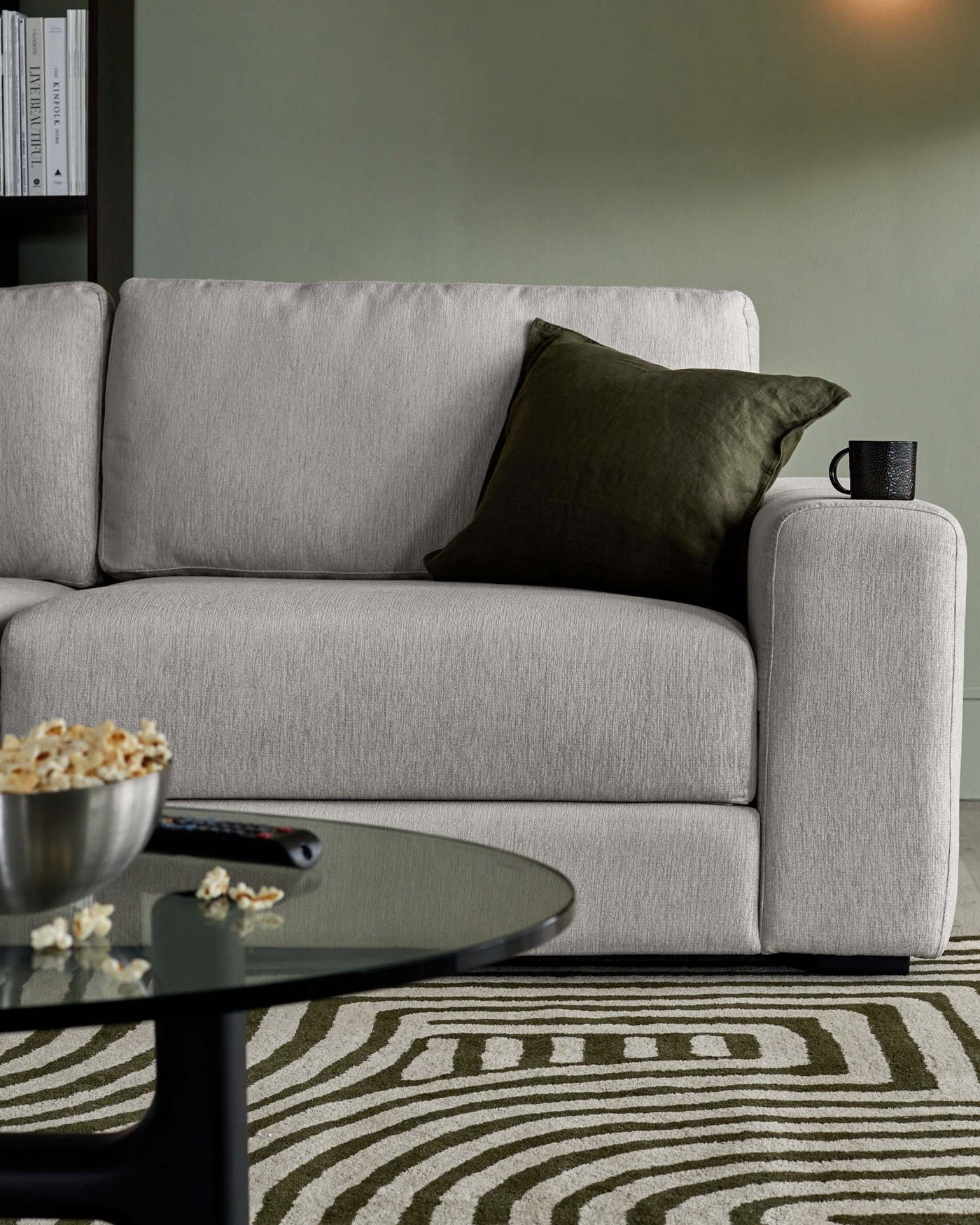 Modern light grey fabric sofa with clean lines and simplistic design, featuring one armrest, complemented with a dark green decorative pillow. In the foreground, there's a round, black coffee table with a glass top, and underneath is a patterned off-white and green area rug. Beside the sofa, a small black cup sits atop a matching narrow cylindrical stand.
