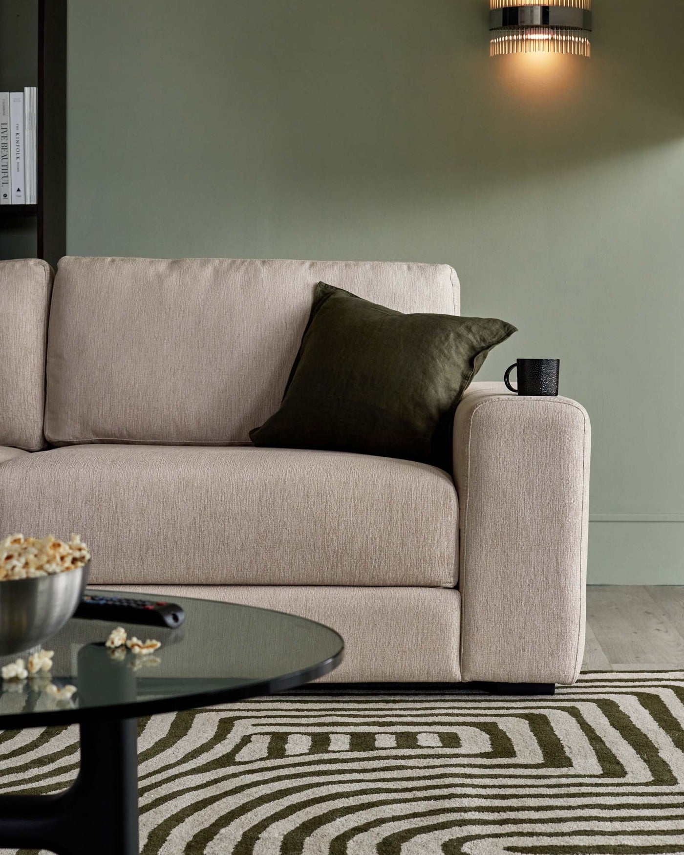 Modern beige fabric sofa with clean lines, featuring plush cushioning, a dark green accent pillow, and a black cylindrical side table topped with a black mug. In the foreground, a round black coffee table presents contrast against a patterned grey and green area rug.