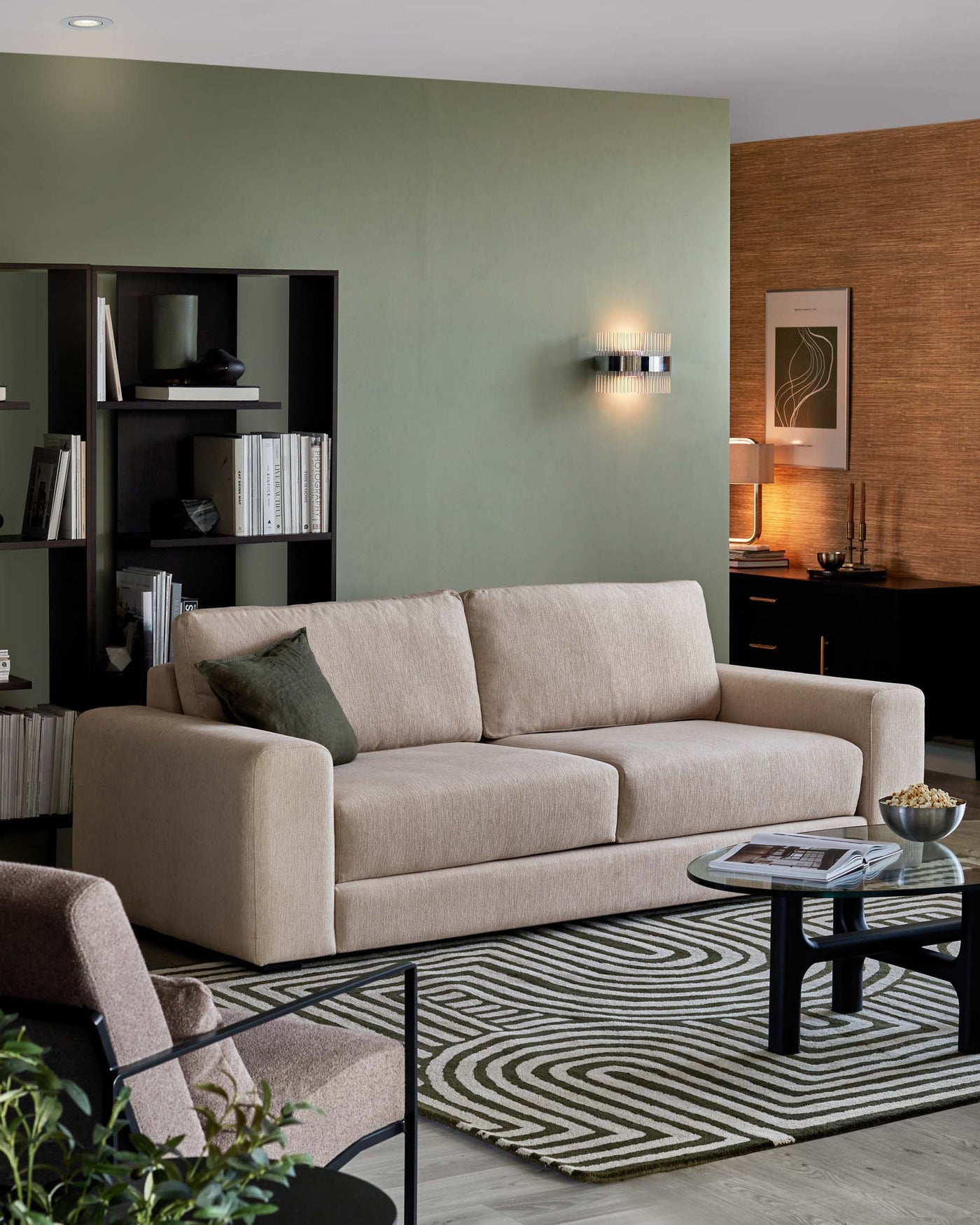Modern living room featuring a neutral-toned three-seater fabric sofa with plush cushions, a black metal-frame coffee table with a round glass top, and a coordinating geometric-patterned area rug in shades of green and beige. A dark-toned bookshelf with various decorative items and books accents the space.