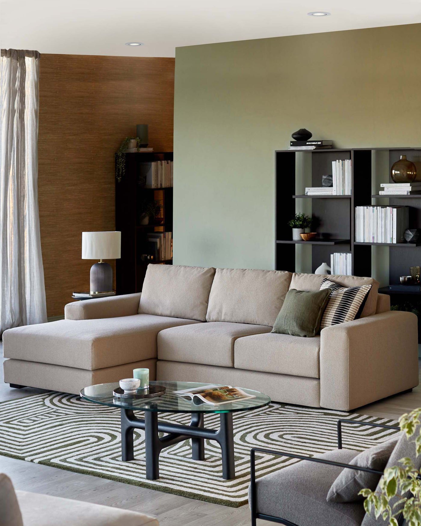 Modern living room showcasing a beige, L-shaped sectional sofa with plush cushions and additional throw pillows in earthy tones. In front of it stands a contemporary round glass-top coffee table with a black frame. To the side, a sleek black bookcase is partially visible, filled with books and decorative items. The furniture is complemented by a patterned area rug with geometric designs, and a matching side table with a lamp is partially seen to the left.