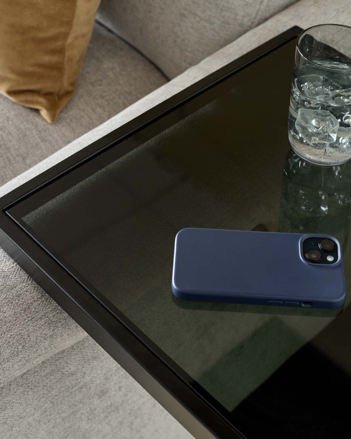 Smoked glass tray table with black frame holding a blue phone case and glass of ice water, styled on Grey sofa.