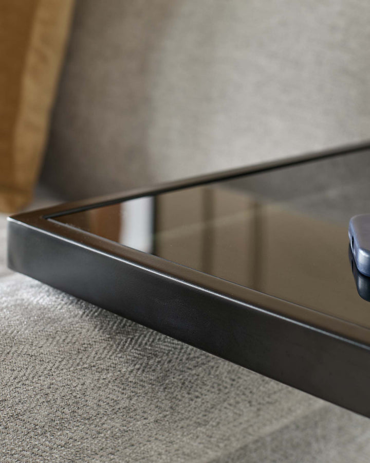 Ezra tray table with smoked glass top and dark metal frame, shown at an angle on light Grey upholstery.