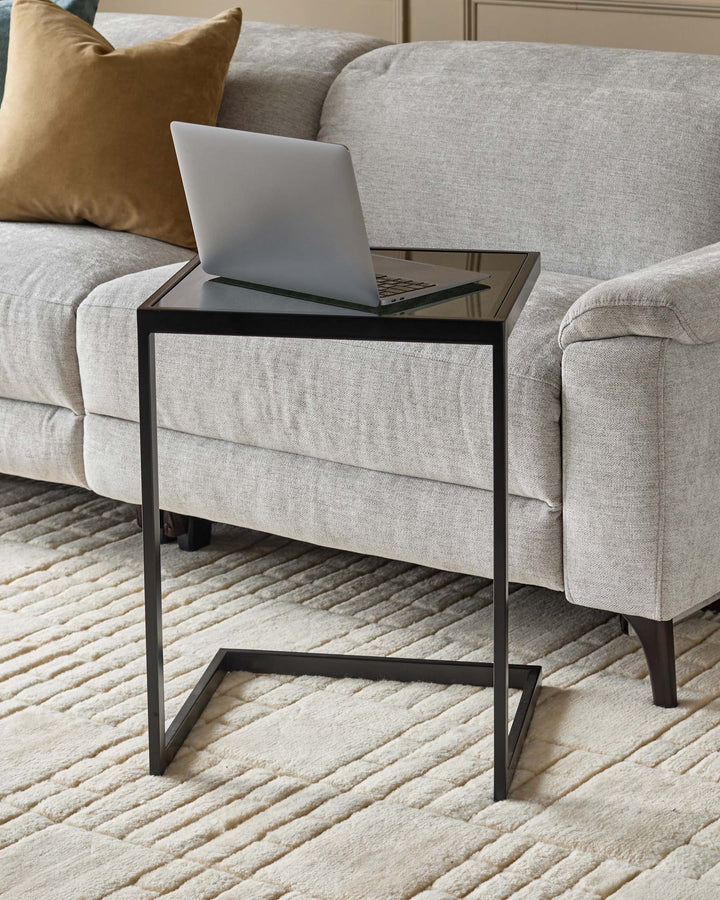 Ezra tray table with smoked glass top and black metal frame, positioned beside a beige sofa with gold pillow.
