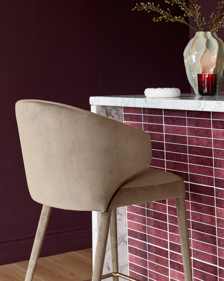 Champagne velvet bar stool with curved backrest and wooden legs, positioned at a marble and burgundy tiled counter.