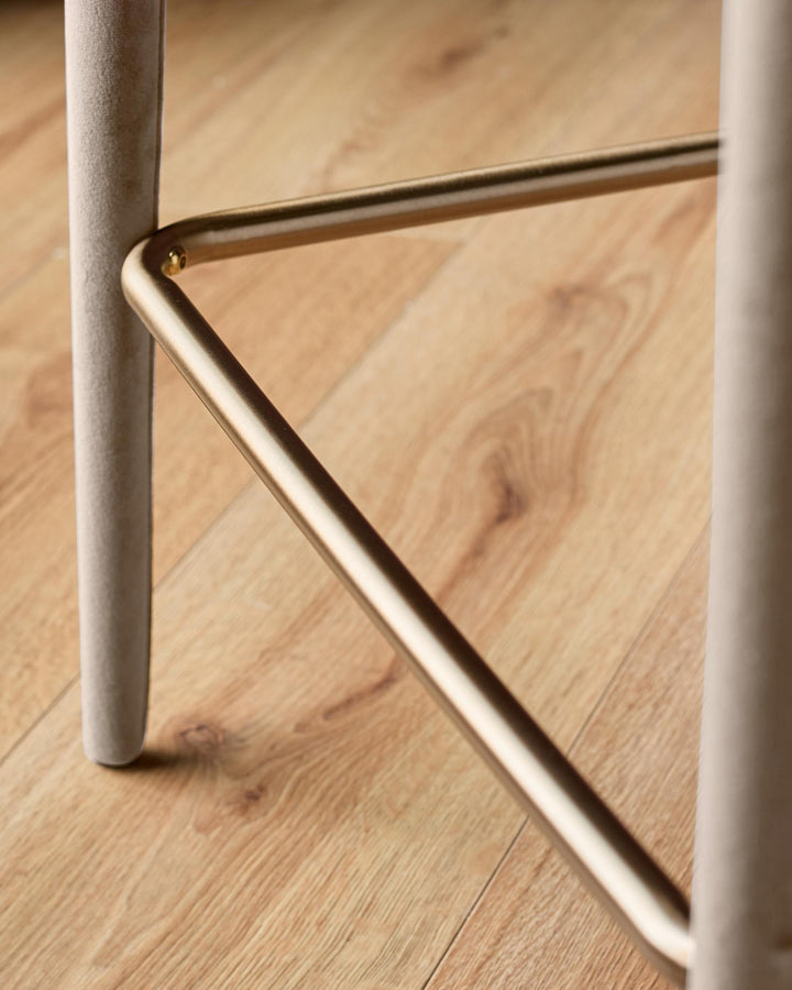 Close-up of champagne velvet bar stool showing brass footrest and white cylindrical leg on wooden floor.